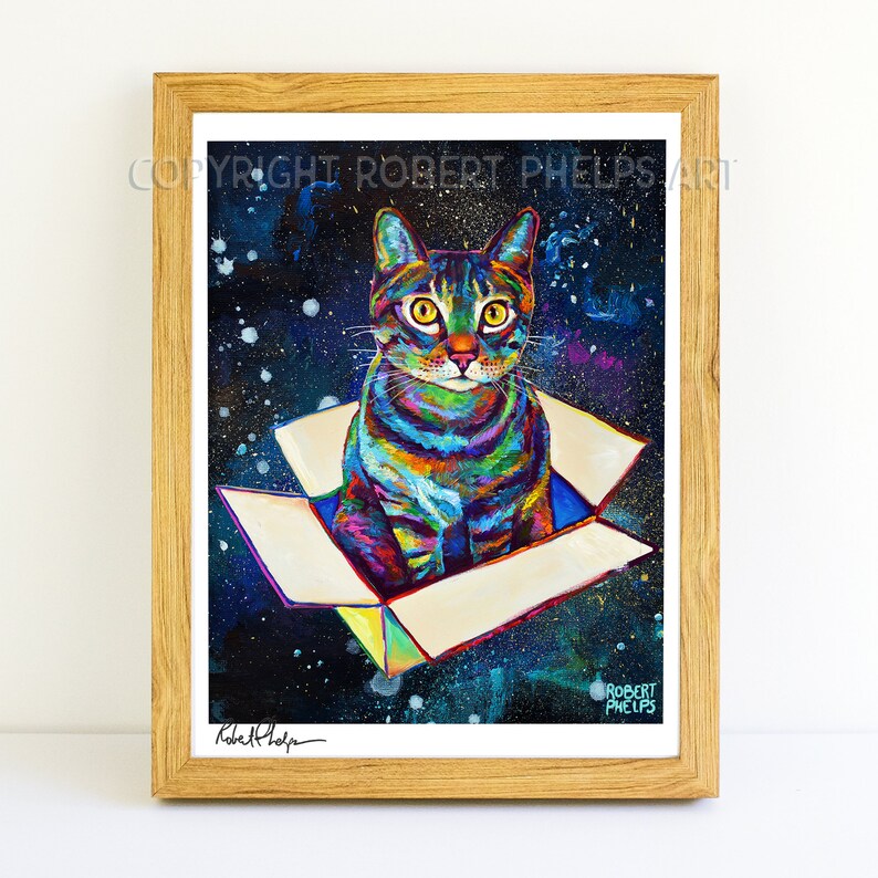 May include: A colorful painting of a cat sitting in a box against a starry night background. The cat is looking directly at the viewer. The painting is framed in a light brown wood frame. The artist's signature is visible in the bottom left corner.