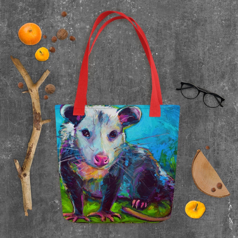 May include: A colorful tote bag featuring a painting of a possum with a blue, green, and purple background. The possum has a white face with black eyes and a pink nose.