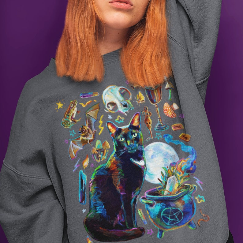 May include: A dark gray sweatshirt with a colorful graphic print of a black cat, a cauldron, and various mystical symbols. The cat is sitting in front of the cauldron, which is filled with a bubbling liquid. The symbols include a skull, a bat, a frog, a crystal, a moon, and a pentagram.