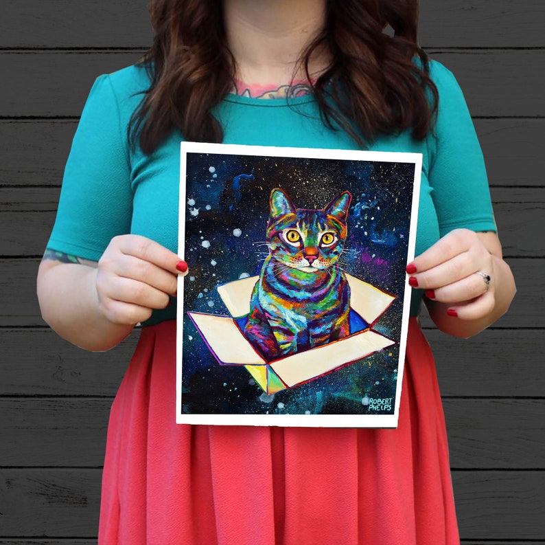 May include: A colorful painting of a cat sitting in a box against a dark blue and purple space background. The cat has green eyes and a striped pattern. The box is white with a brown interior. The painting is signed "Robert Phelps".