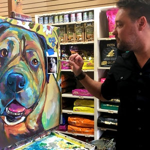 May include: A colorful painting of a dog's face on a canvas easel. The artist is using a brush to add details to the painting. The painting is in a pet store with shelves of pet food in the background.