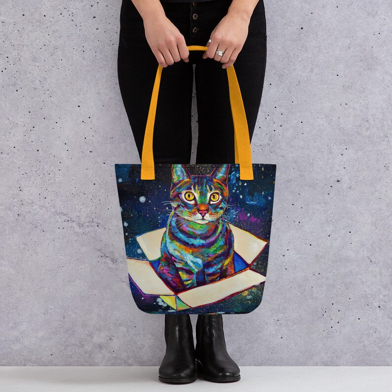 May include: A tote bag with yellow handles features a colorful cat sitting inside a box against a dark blue galaxy background.