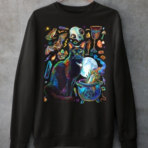 May include: Black sweatshirt with a colorful illustration of a black cat, a cauldron, and other witchy items. The cat is sitting in front of the cauldron, which is filled with a green liquid. The illustration is surrounded by stars and other magical symbols.