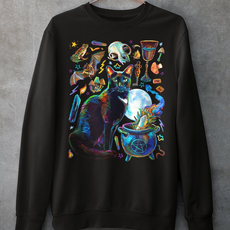 May include: Black sweatshirt with a colorful illustration of a black cat, a cauldron, and other witchy items. The cat is sitting in front of the cauldron, which is filled with a green liquid. The illustration is surrounded by stars and other magical symbols.