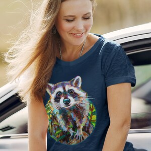 May include: A navy blue t-shirt with a colorful illustration of a raccoon in a circle. The raccoon is looking at the viewer with a curious expression.