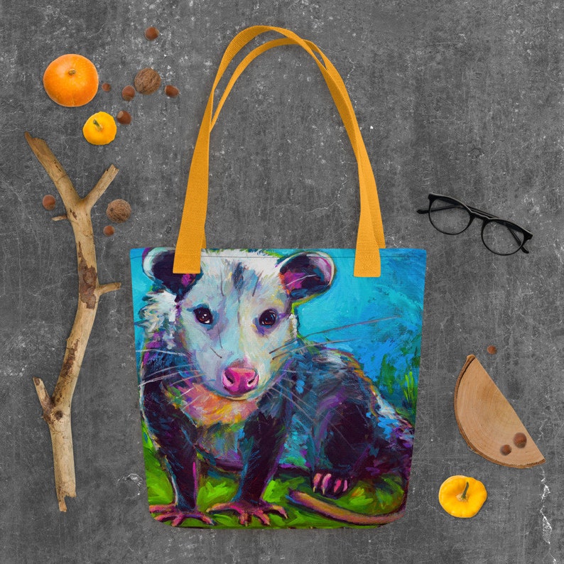 May include: A colorful tote bag featuring a close-up painting of a possum with a bright blue background. The possum has a white face with black eyes and a pink nose. The bag has yellow straps.