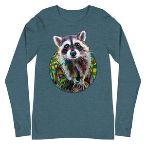 May include: Teal long-sleeve shirt featuring a colorful raccoon illustration within a circular frame. The raccoon has a detailed, artistic design with shades of brown, white, and black. The background includes green and yellow foliage.