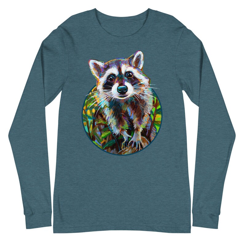 May include: Teal long-sleeve shirt featuring a colorful raccoon illustration within a circular frame. The raccoon has a detailed, artistic design with shades of brown, white, and black. The background includes green and yellow foliage.
