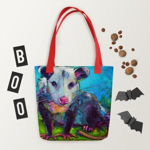 May include: A red tote bag with a colorful painting of a possum on the front. The possum is sitting on a green background with a blue sky behind it. The possum has a white face with black eyes and a pink nose.