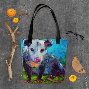 May include: A tote bag with a colorful painting of a possum on a blue background. The possum is looking directly at the viewer with a curious expression. The bag has black straps.