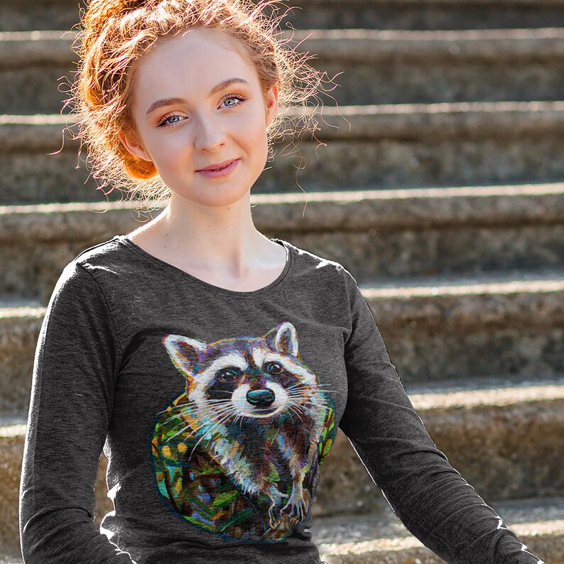 May include: Dark gray long-sleeve shirt featuring a colorful raccoon graphic. The raccoon has a detailed, artistic design with a variety of colors. The shirt has a scoop neck and long sleeves. The background is a set of concrete steps.