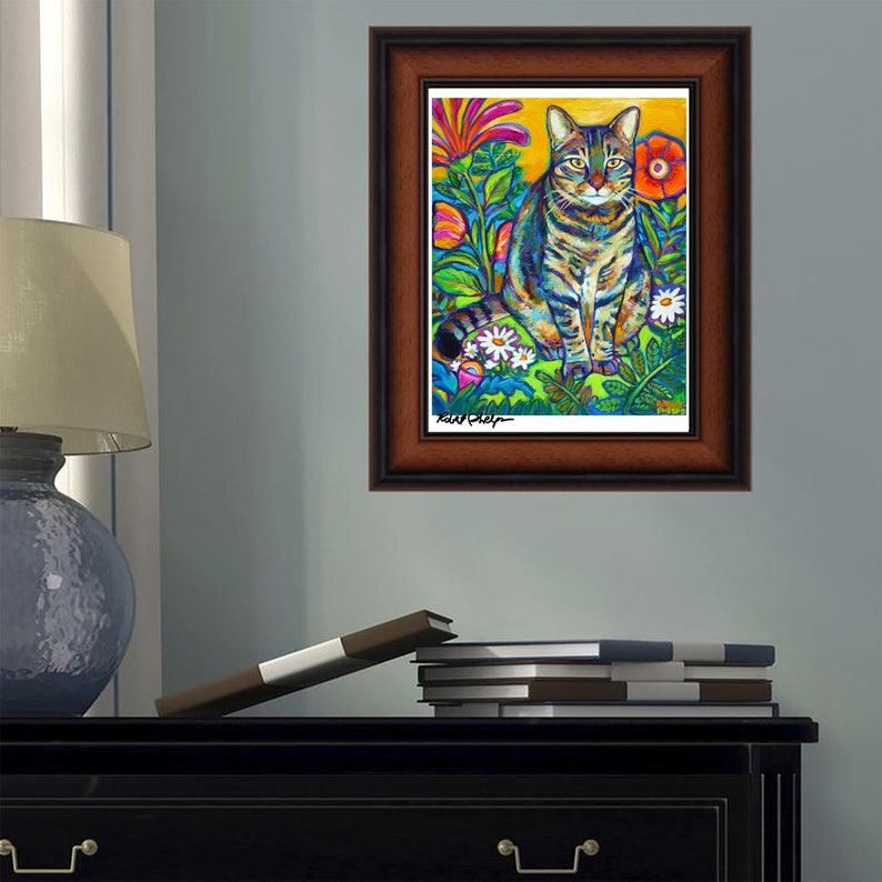 May include: A framed painting of a tabby cat sitting in a field of colorful flowers. The cat has green eyes and a striped pattern. The flowers are pink, yellow, and white. The painting is signed "Ken Daley".