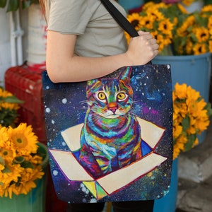 May include: A colorful tote bag with a galaxy background and a cat sitting in a box. The cat is painted in a rainbow of colors and has a surprised expression.