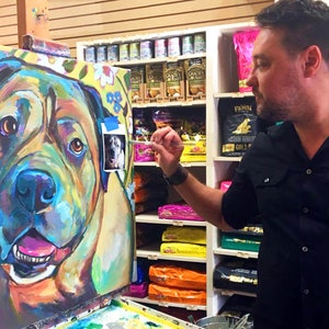 May include: A colorful painting of a dog's head on a canvas easel. The artist is painting the dog's nose with a brush. The painting is in a pet store with shelves of pet food in the background.