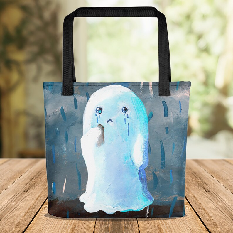 May include: A tote bag featuring a sad, light blue ghost with tears and a tissue in a rainy scene. The bag has black handles and a dark blue background with vertical blue streaks.