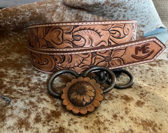 May include: A light brown leather belt with floral carvings and a decorative buckle. The belt features detailed embossed flowers and foliage. The buckle has a sunflower design and is connected to the belt with metal rings. The belt's end has the initials "ME".
