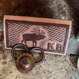 May include: A light brown leather wallet with a woven pattern and an elk silhouette. The wallet is personalized with the initials "KB".
