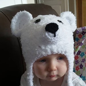 May include: A white crocheted polar bear hat with black eyes and nose. The hat has long white tassels. A child is wearing the hat.