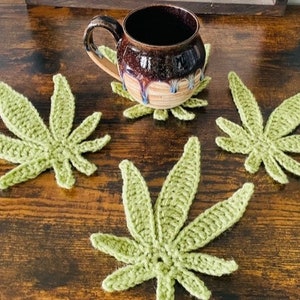 May include: Four crocheted green marijuana leaf coasters on a wooden surface. A brown ceramic mug sits on one of the coasters.