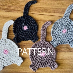 May include: Four crocheted cat butt coasters in different colors: white, black, gray, and brown. Each coaster has a pink circle in the center.