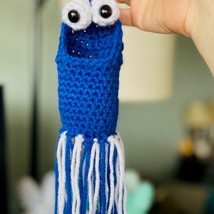 Op de afbeelding: Een blauwe gehaakte octopus met witte tentakels en goggle-ogen. De octopus hangt aan een lus van garen.