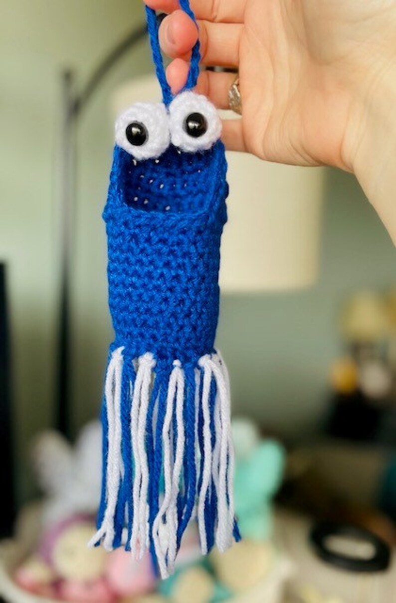 Op de afbeelding: Een blauwe gehaakte octopus met witte tentakels en goggle-ogen. De octopus hangt aan een lus van garen.