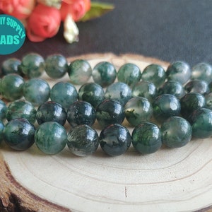 May include: A strand of green moss agate beads, each bead is round and has a mottled green and white pattern. The beads are arranged in a circle on a wooden surface.