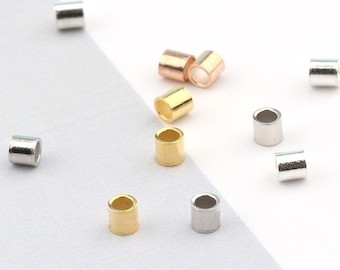 May include: Small, cylindrical beads in gold, silver, and rose gold tones are scattered across a white surface. The beads are made of metal and have a smooth, polished finish. The image includes a teal circle with the text "ANNY DIY SUPPLY BEADS".