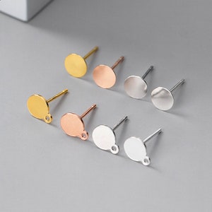 May include: An assortment of metal earring posts in gold, rose gold, and silver. Each post has a flat, circular disc and a small loop for attaching earring components. The background is a light grey surface.