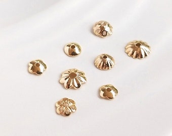 Può includere: Assortimento di cappucci per perline in metallo dorato in varie forme e dimensioni, tra cui design rotondi, floreali e a festoni. I cappucci per perline sono esposti su una superficie bianca.