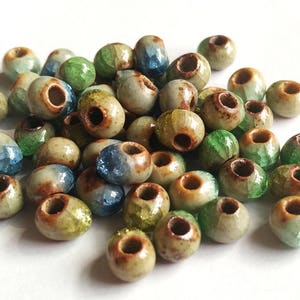 May include: A pile of ceramic beads in various shades of green, blue, and brown. The beads have a speckled glaze and a hole in the centre for stringing.