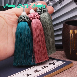 May include: Three tassel ornaments with a knotted top and a white bead. The tassels are teal, pink, and gray.