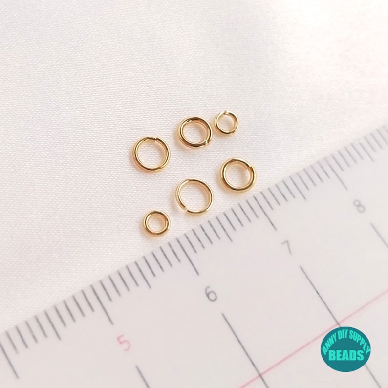 May include: A set of six gold jump rings of varying sizes, measured against a ruler with markings in centimeters. The jump rings are used for jewelry making and crafting.