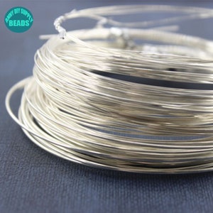 Peut inclure: Gros plan sur une bobine de fil argent&eacute;, &eacute;troitement enroul&eacute;. Le fil est fin et semble souple, id&eacute;al pour l'artisanat ou la bijouterie. L'image comprend le texte "ANNY DIY SUPPLY BEADS" dans un cercle turquoise.