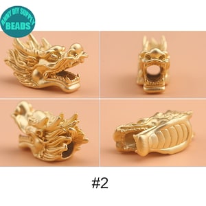 May include: Gold-colored dragon head charm with intricate details. The charm has a hollow center and is designed to be used as a bead or pendant.