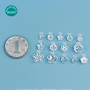 May include: A collection of silver-colored flower-shaped beads, each with a unique design, displayed on a light blue surface. A coin is placed next to the beads for size comparison. The image includes the text "ANNY DIY SUPPLY BEADS".