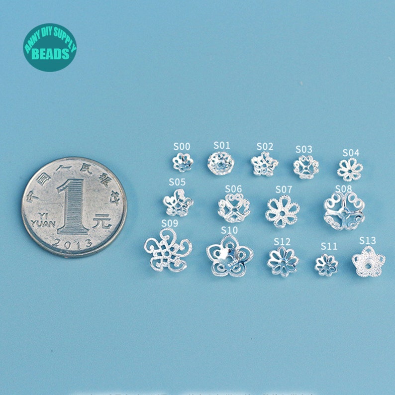 May include: A collection of silver-colored flower-shaped beads, each with a unique design, displayed on a light blue surface. A coin is placed next to the beads for size comparison. The image includes the text "ANNY DIY SUPPLY BEADS".