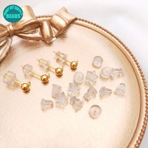 Puede incluir: Primer plano de un conjunto de respaldos para pendientes de diferentes formas y tamaños. Los respaldos están hechos de plástico transparente y están diseñados para ajustarse sobre el poste de un pendiente. También hay tres postes de pendientes de oro con pequeñas bolas de oro en el extremo.