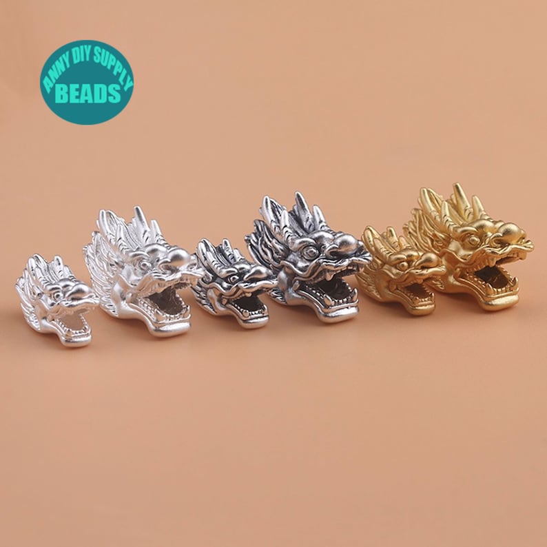 May include: Five silver and gold dragon head beads. The beads are in different sizes and are arranged in a row from smallest to largest. The beads are all facing the right side of the image.