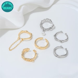 May include: A collection of five open rings in gold and silver tones. One gold ring features a delicate chain attachment, another gold ring has a textured surface, and one silver ring includes a small white pearl charm. Two silver rings are plain.