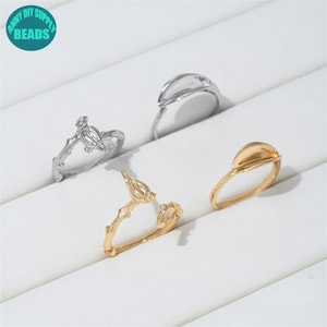Puede incluir: Cuatro anillos de metal dispuestos en grupo. Dos anillos presentan un diseño de rama texturizada con una pequeña figura de pájaro, uno en tono plateado y otro en dorado. Dos anillos presentan un elemento liso y curvo en forma de hoja, en plata y oro.