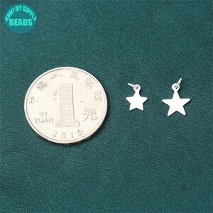 May include: Two silver star charms with jump rings. The charms are on a green fabric background with a Chinese yuan coin.