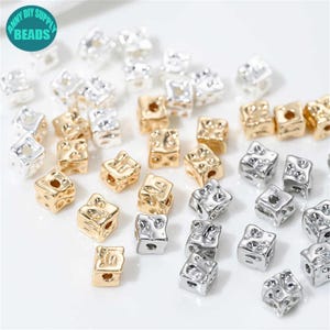 Peut inclure: Des perles cubiques métalliques dorées et argentées, à surface texturée, dispersées sur un fond blanc. L'image comprend un logo indiquant "ANNY DIY SUPPLY BEADS".