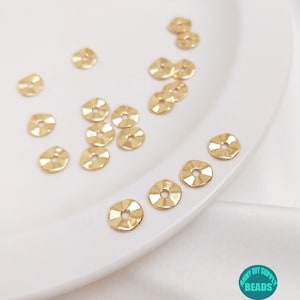 May include: A close-up of a white plate with 15 gold-coloured metal spacer beads. The beads are round with a slightly raised centre and a hole in the middle. The beads are arranged in a random pattern on the plate.