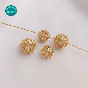 May include: Four gold-toned metal beads with a filigree design and clear crystal accents. The beads are arranged on a white surface.