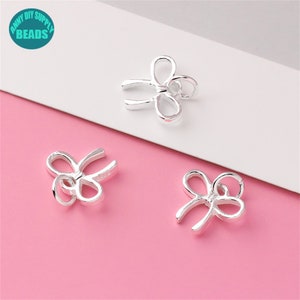 May include: Three silver-colored bow-shaped charms are displayed against a white and pink background. The charms have a polished finish and are designed with looped ends, suggesting they are jewelry components or decorative elements. The charms are from ANNY DIY SUPPLY BEADS.