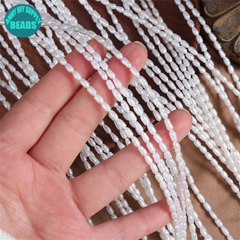 K&ouml;nnte beinhalten: Nahaufnahme von mehreren Str&auml;ngen wei&szlig;er, oval geformter Perlen. Die Perlen sind aufgef&auml;delt und erzeugen ein flie&szlig;endes, strukturiertes Aussehen. Eine Hand h&auml;lt einen Teil der Perlen, der Rest f&auml;llt kaskadenartig nach unten. Das Bild enth&auml;lt ein Logo mit der Aufschrift "ANNY DIY SUPPLY BEADS".