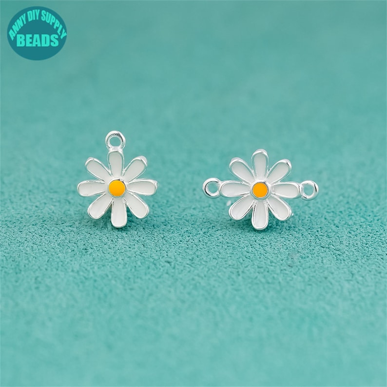 K&ouml;nnte beinhalten: Zwei charmartige Anh&auml;nger in Form von G&auml;nsebl&uuml;mchen mit wei&szlig;en Bl&uuml;tenbl&auml;ttern, gelben Zentren und silberfarbenen Metallr&auml;ndern. Jeder Anh&auml;nger hat eine Schlaufe zum Befestigen an Schmuck. Der Hintergrund ist einfarbig t&uuml;rkis, und der Text "ANNY DIY SUPPLY BEADS" befindet sich in einem Kreis oben links.