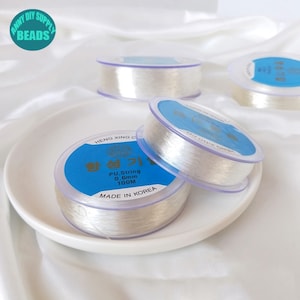 以下が含まれることがあります： ジュエリー作りのための透明な伸縮性のある糸の糸巻きが3つ。糸巻きには「PU String 0.6mm 100M Made in Korea」とラベルが貼られています。