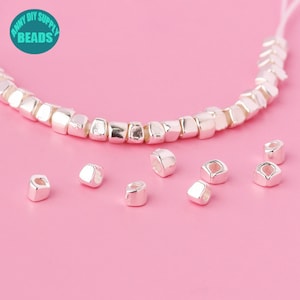 Può includere: Perle quadrate argentate con un foro al centro. Le perle sono disposte in fila e sparse su una superficie rosa. Le perle sono per la creazione di gioielli.